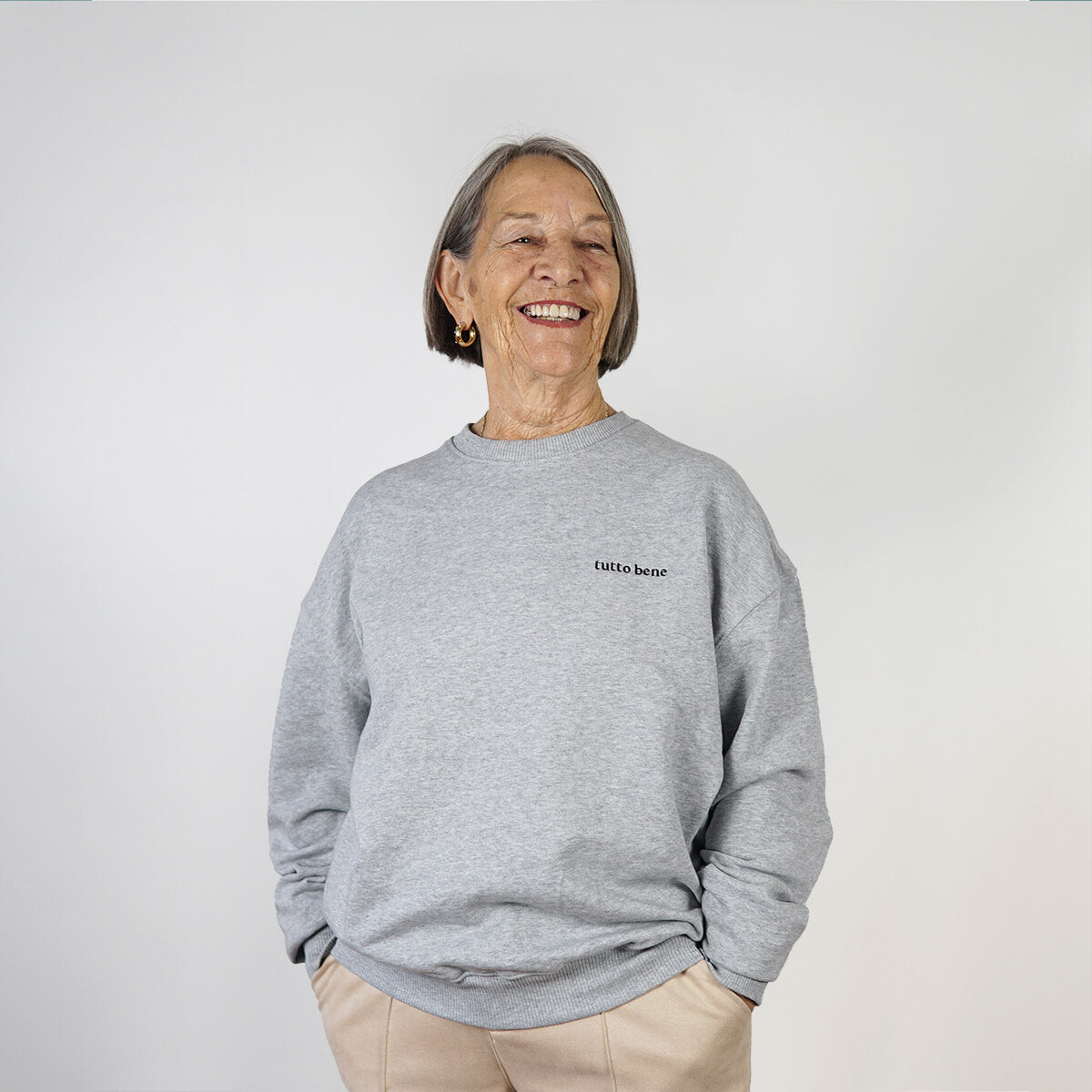 Ältere Frau die einen grauer Sweater der Marke "studio ciao" mit "tutto bene" Stickerei auf der Brust trägt.